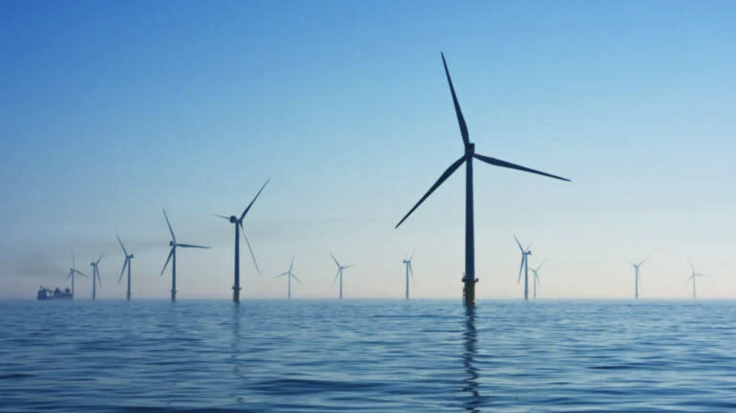A row of wind turbines stands in the ocean, with their blades rotating in the breeze. The scene is set against a clear blue sky, reflecting on the calm water